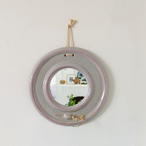 May include: A round, grey ceramic mirror with a pink rim and a rope hanger. The mirror is hanging on a white wall above a light blue dresser.