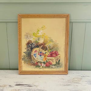 Peut inclure: Une peinture vintage encadrée représente un panier débordant de fruits et de fleurs colorés. L'œuvre d'art est encadrée dans du bois et se détache sur un fond jaune clair. Le tableau est posé sur un meuble blanc.