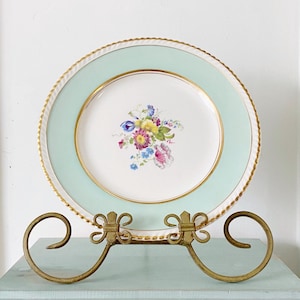 Peut inclure: Une assiette vintage à motif floral avec un bord doré et une bordure bleu clair est exposée sur un support en métal doré. Le support est sur une table en bois peinte en bleu clair avec un tiroir.