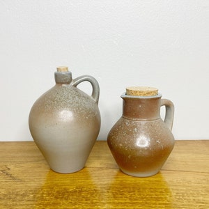 Peut inclure: Deux cruches en céramique avec des bouchons en liège. La plus grande cruche est de couleur gris clair avec un glaçage moucheté. La plus petite cruche est de couleur gris foncé avec un glaçage moucheté brun. Les deux cruches ont une poignée sur le côté.