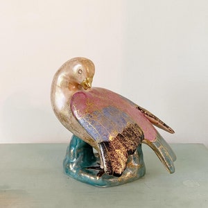 May include: A ceramic dove figurine with a pink, blue, and gold painted design. The dove is perched on a teal base.