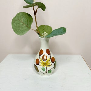 Peut inclure: Un vase en céramique blanche avec un motif floral dans les tons de brun, jaune et vert. Le vase est rempli d'une branche d'eucalyptus.