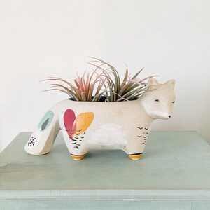 Peut inclure: Planteur en céramique blanche en forme de renard avec des motifs géométriques colorés, contenant une petite plante aérienne verte et rose. Le planteur est posé sur une table en bois bleu clair avec un tiroir.