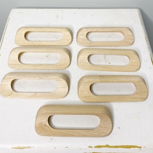 Peut inclure: Huit poignées de tiroir en bois non fini de différentes tailles et formes, disposées sur une surface blanche.