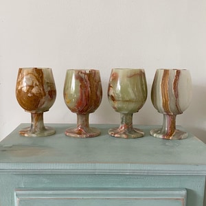 Puede incluir: Cuatro copas de vino decorativas de ónix con un patrón veteado único en tonos marrón, verde y rojo. Las copas se exhiben sobre un mueble azul claro. Cada copa tiene un tallo y una copa redondeada.