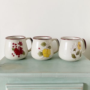 Peut inclure: Trois tasses en céramique blanche avec des bords marrons et des motifs floraux. Les tasses ont des motifs floraux différents, y compris des fleurs rouges et vertes, des fleurs jaunes et des fleurs blanches avec des feuilles vertes.
