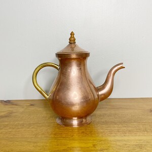 Peut inclure: Une théière en cuivre avec une poignée en laiton et un bouton en bois sur le couvercle. La théière est posée sur une surface en bois.