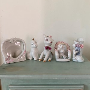 Peut inclure: Cinq figurines en porcelaine sur une surface en bois bleu clair. Les figurines sont blanches avec des accents roses et dorés. Une figurine est un cadre en forme de cœur, une autre est une licorne, une autre est un cheval avec un nœud rose, une autre est un chat assis et la dernière est un cerf avec un nœud rose.