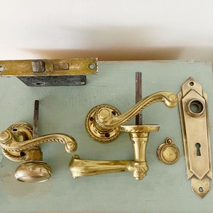 May include: A set of antique brass door hardware, including a door knob, a door handle, a door plate, and a door latch. The hardware is displayed on a light blue surface.