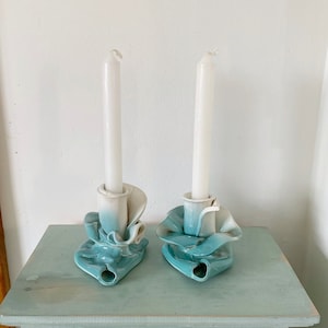 May include: Two white candles in blue and white ceramic candle holders with a ruffled design. The candle holders are on a light blue wooden table with a drawer.