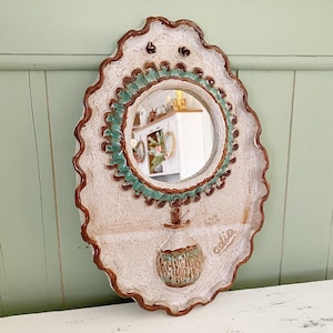 May include: Oval ceramic mirror with a white, textured surface and a scalloped edge. The mirror has a round centre with a turquoise border and a small vase design below. The word "adia" is written on the right side.