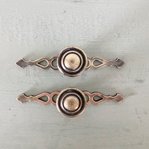 Peut inclure: Deux poignées de meuble de style antique avec une finition vieillie. Chaque poignée présente un bouton central circulaire avec des anneaux concentriques, flanqué de poignées décoratives et allongées. La quincaillerie est de couleur laiton mat, sur une surface bleu-vert pâle.