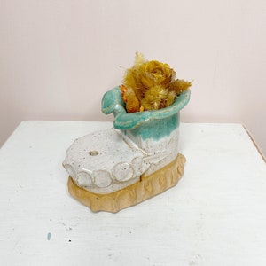 Peut inclure: Un pot en céramique en forme de botte avec un glaçage blanc et turquoise. Le pot est rempli de fleurs séchées.