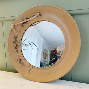 May include: Circular, beige ceramic mirror featuring a floral pattern and a natural rope for hanging. The frame has a thick, rounded edge. The reflection shows a room with a Christmas tree. A unique piece for home decor.