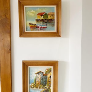 Peut inclure: Deux tableaux encadrés sur le thème nautique. Le tableau du haut représente un petit village avec des bateaux dans l'eau. Le tableau du bas représente un bâtiment en pierre avec un toit rouge et un jardin.