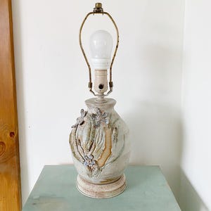 May include: A ceramic table lamp with a white and brown glaze. The lamp features a floral design with raised details. The lamp has a brass base and a white light bulb.