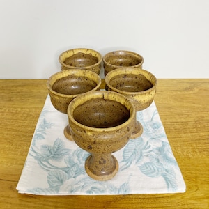 May include: Set of five brown and yellow speckled ceramic cups with a footed base. The cups are arranged in a circle on a white and blue floral tablecloth.