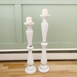 Peut inclure: Deux bougeoirs en bois blanc avec une finition vieillie. Les bougeoirs sont hauts et fins, avec une base ronde et un sommet évasé. Chaque bougeoir a une bougie blanche sur le dessus.