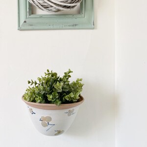 Peut inclure: Un pot de fleurs suspendu en céramique blanche avec un motif floral et une plante verte à l'intérieur. Le pot est suspendu à un mur blanc.
