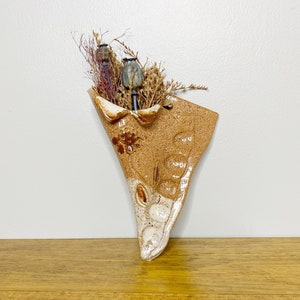 Peut inclure: Un vase mural en céramique brun et blanc avec une forme asymétrique unique. Le vase est rempli de fleurs séchées et de gousses de graines.