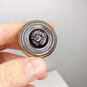 Puede incluir: Un tapón de botella de plata y oro con un diseño floral. El tapón tiene un centro negro con un diseño floral plateado.