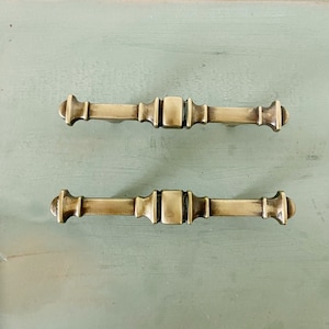 May include: Two antique brass cabinet pulls with a square centre and a decorative design on the ends. The pulls are on a light green surface.