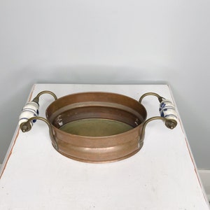 Peut inclure: Un bol de service en cuivre avec deux poignées en céramique blanche ornées de motifs floraux bleus. Le bol a une finition martelée et repose sur une surface blanche.