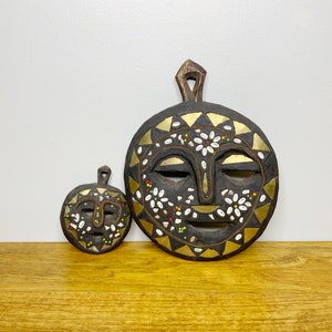 Peut inclure: Deux masques en bois avec un design de visage humain. Le plus grand masque est décoré d'accents blancs et dorés, tandis que le plus petit masque est décoré d'accents blancs et noirs.