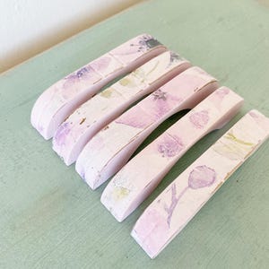 Peut inclure: Cinq poignées de tiroir en bois rose avec un motif floral violet et blanc. Elles sont disposées sur une surface vert clair. Chaque poignée présente un motif floral unique et une finition vieillie, pour un style vintage.