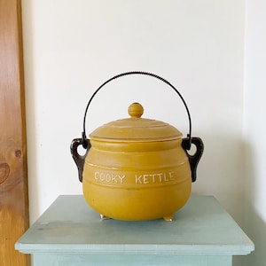 Peut inclure: Une bouilloire en céramique jaune avec un couvercle et une poignée noire. La bouilloire a une forme arrondie et repose sur une table bleu clair. Le texte "COOKY KETTLE" est en relief sur le devant.