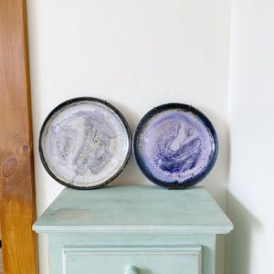 May include: Two ceramic plates with a white glaze and purple and blue speckles. The plates are hanging on a light blue wall above a light blue dresser with a single drawer.