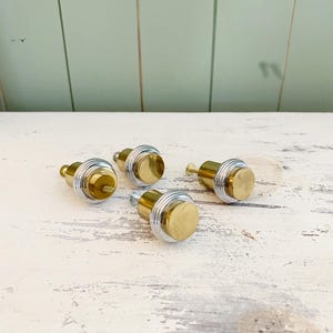 Peut inclure: Quatre boutons de meuble en laiton et argent sont disposés sur une surface en bois blanc vieilli. Chaque bouton présente une face circulaire en laiton et un corps argenté strié. Les boutons sont conçus pour les meubles ou les armoires.