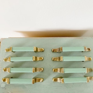 May include: Eight light green cabinet knobs with gold metal accents. The knobs are arranged in two rows of four on a light green surface.