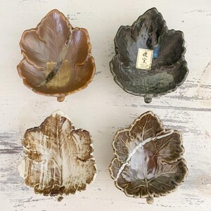 Peut inclure: Quatre coupelles en céramique en forme de feuilles d'érable. L'une est brun clair, une autre gris anthracite avec une étiquette "Made in Japan", une troisième crème avec des coups de pinceau bruns, et la dernière brun brillant avec un motif craquelé.