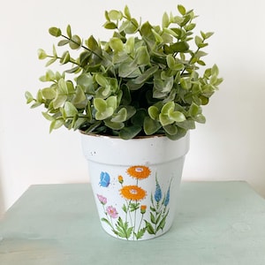 Peut inclure: Un pot en céramique blanche avec un motif floral composé de fleurs orange, roses et bleues et un papillon bleu. Le pot est rempli de plantes vertes feuillues.