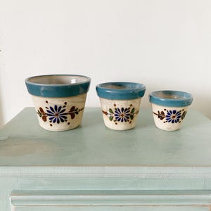 Puede incluir: Tres macetas de cerámica con bordes azules y diseños florales en tonos azules, marrones y blancos. Las macetas están ordenadas por tamaño, con la maceta más grande a la izquierda y la más pequeña a la derecha.