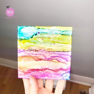 May include: A small square canvas painting with abstract layers of blue, green, yellow, orange, and pink paint.