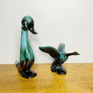 以下が含まれることがあります： 緑と茶色の釉薬をかけたセラミック製の鴨の置物2つ。大きな鴨は頭を横に傾けて直立している。小さな鴨は羽を広げて飛んでいる。