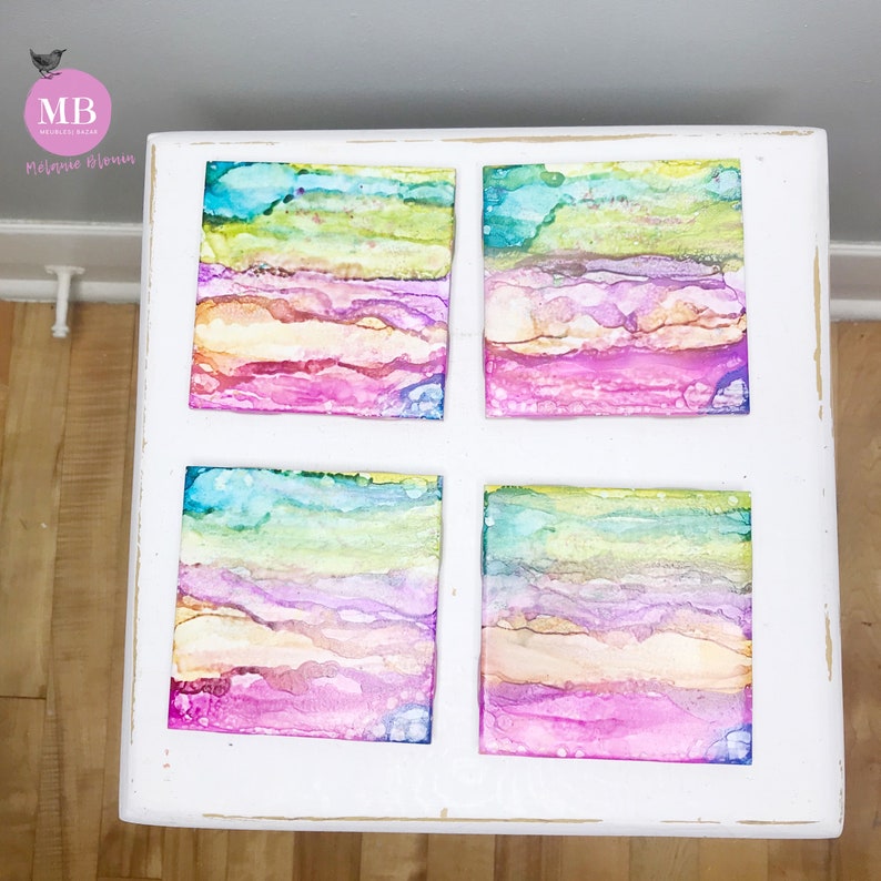 May include: Four square coasters with abstract watercolor designs in shades of pink, blue, green, and yellow. The coasters are arranged in a grid pattern on a white wooden surface.