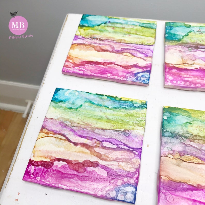 May include: Four square coasters with a colorful abstract design featuring shades of pink, blue, green, and yellow. The coasters have a layered, marbled effect.