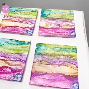 May include: Set of four square coasters with abstract art. The coasters feature a layered design with shades of pink, purple, green, blue, and yellow.