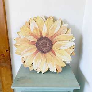 Peut inclure: Une grande décoration de tournesol en papier avec des pétales jaunes et un centre brun. Le tournesol repose sur une table de chevet bleu clair avec un seul tiroir et un bouton rond.
