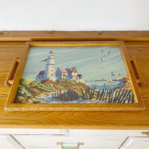 May include: A framed paint-by-numbers picture of a coastal scene featuring a lighthouse, a house, and a sailboat. The artwork is in a wooden frame with handles. The colour scheme includes blues, greens, and browns, creating a vintage aesthetic.