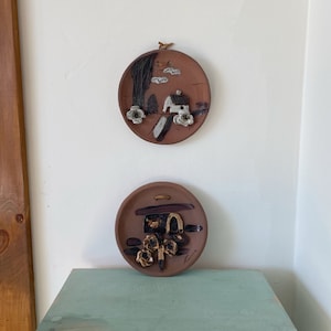Peut inclure: Deux plaques murales rondes en céramique avec des motifs en relief 3D. La plaque du haut présente un paysage brun et blanc avec une petite maison blanche et des fleurs blanches. La plaque du bas présente un motif abstrait brun et noir avec des fleurs blanches.