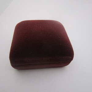 May include: A maroon velvet jewelry box. The box is square with rounded edges and a hinged lid. The velvet material has a soft, textured appearance. The box is likely designed to hold rings, earrings, or other small jewelry items.
