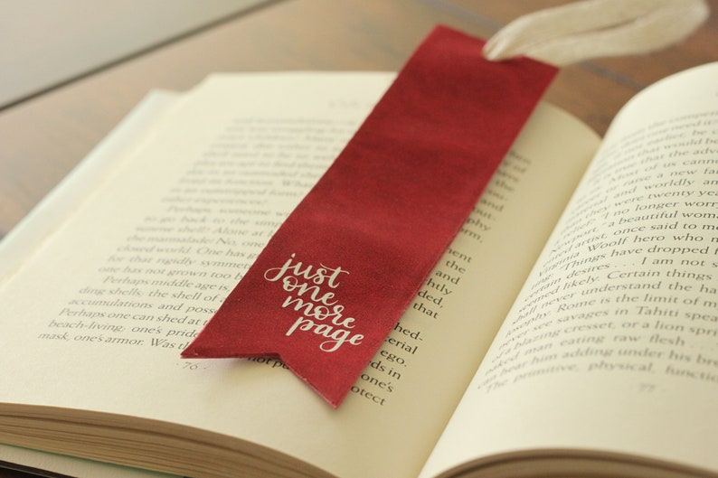 May include: A red bookmark with the text "just one more page" in white script is placed in an open book. The bookmark has a white ribbon attached to the top.