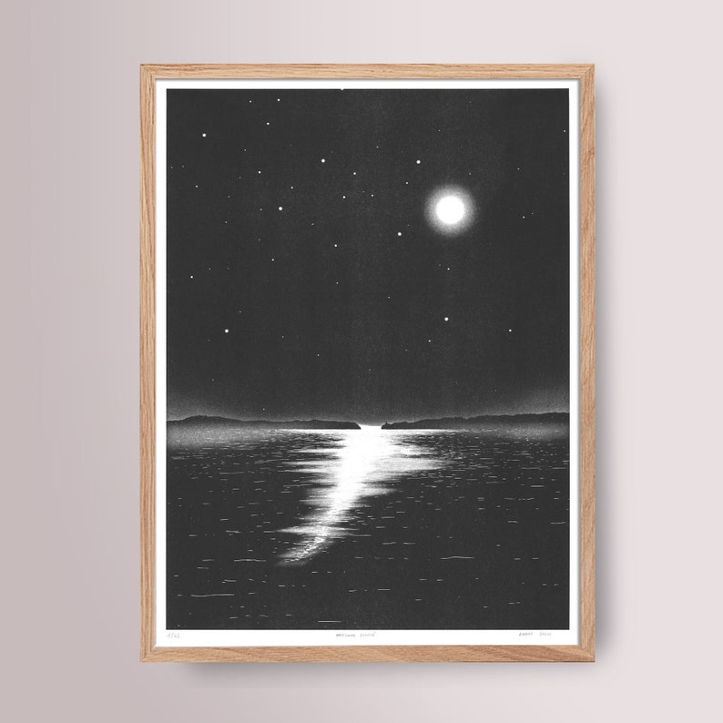 May include: Monochrome print of a moonlit seascape, framed in natural wood. The artwork shows a bright full moon and stars above calm water, with the moon's reflection creating a shimmering path. The print is signed and numbered.