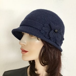 May include: Navy blue cloche hat with a small brim and a decorative flower on the side. The flower is made of the same material as the hat and features a black button center. This hat is a stylish accessory.