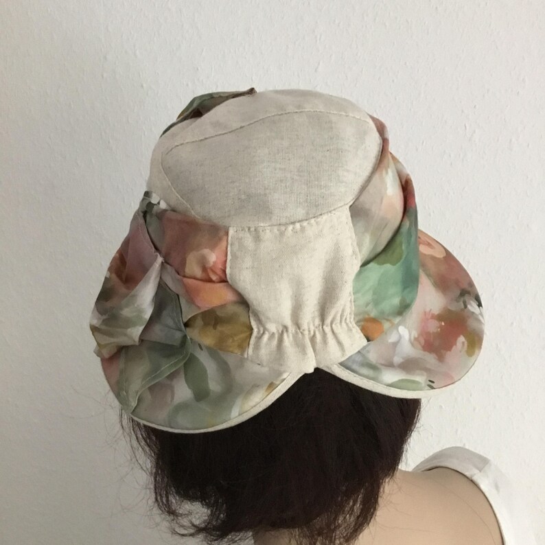 May include: A beige and floral print hat with a brim. The hat features a beige crown and a brim with a colorful floral pattern in shades of pink, green, and orange. The hat is designed for sun protection.
