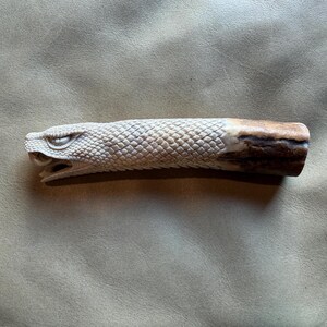 May include: A carved antler handle featuring a detailed snake head at one end. The snake head has visible scales and an open mouth, transitioning into a textured, scale-like pattern along the body, ending in a smooth, brown, natural antler base.